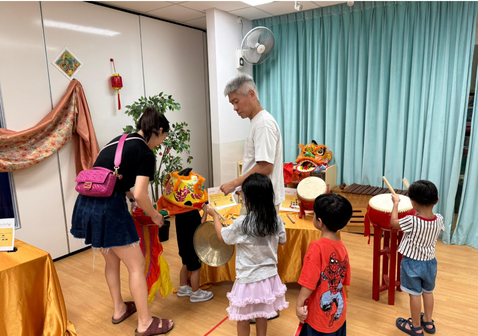 Adults and children try lion dance props and drums in a room with blue curtains and gold-covered tables.